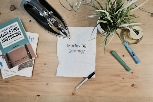 Piece of paper with marketing strategy written on surrounded by pencils, a plant and marketing books