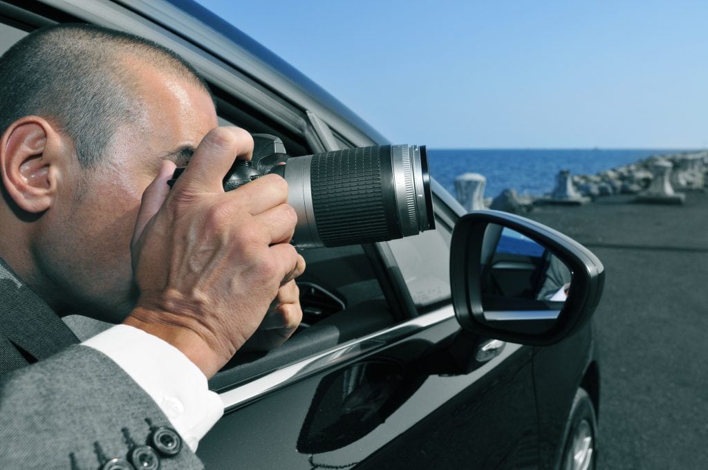 A private investigator taking pictures leaning outside his car. 