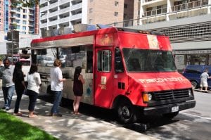 Red food truck with a logo at the front. A long line of people waiting to get food out of a food truck business. He has done a good job promoting his food truck 