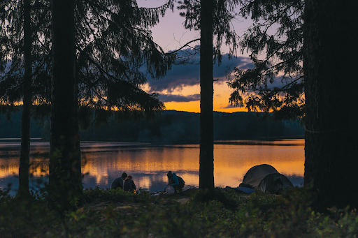 Group of people camping in front of a lake surrounded by trees with a sunset over the mountainscape in the background. The colors, shapes, and elements in this photo encapsulate what outdoorsy people love.