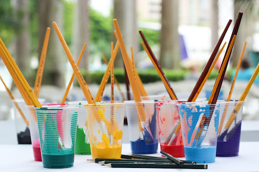 Several plastic cups filled with different colors of paint that contain paintbrushes