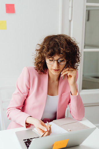 Professional businesswoman in pink blazer answers a phone call while working with feminine logos at desk.