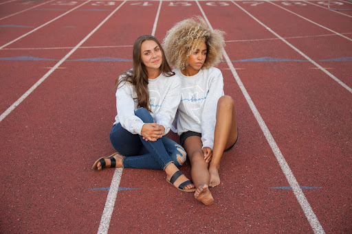 Two girls sitting on a running track wearing matching Portland Gear® sweaters which is an artful logo indeed.