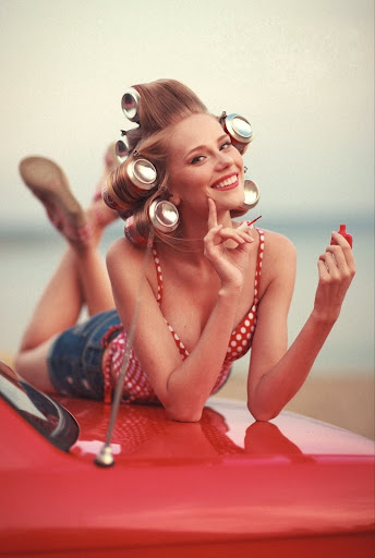 Pin up girl laying on a red car smiling to the camera while doing her nails
