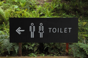  A dark sign positioned in front of a cluster of plants featuring the universal restroom symbol depicting a male and female figure with an arrow pointing to the left