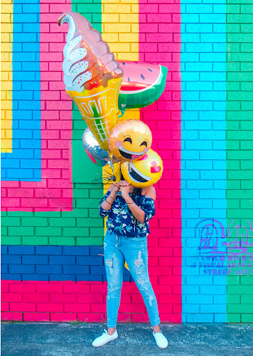 A person covering his face with balloons in the background there is a very colourful wall