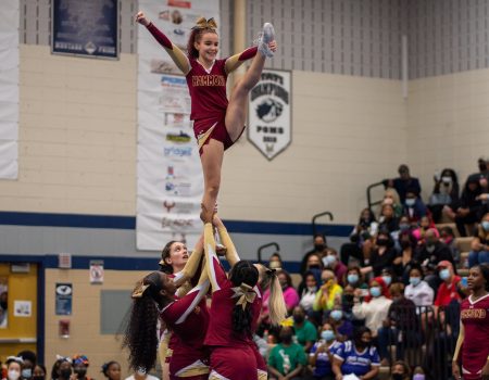 Cheerleader standing with one leg high up being supported by her squad. Sparkling cheer logo